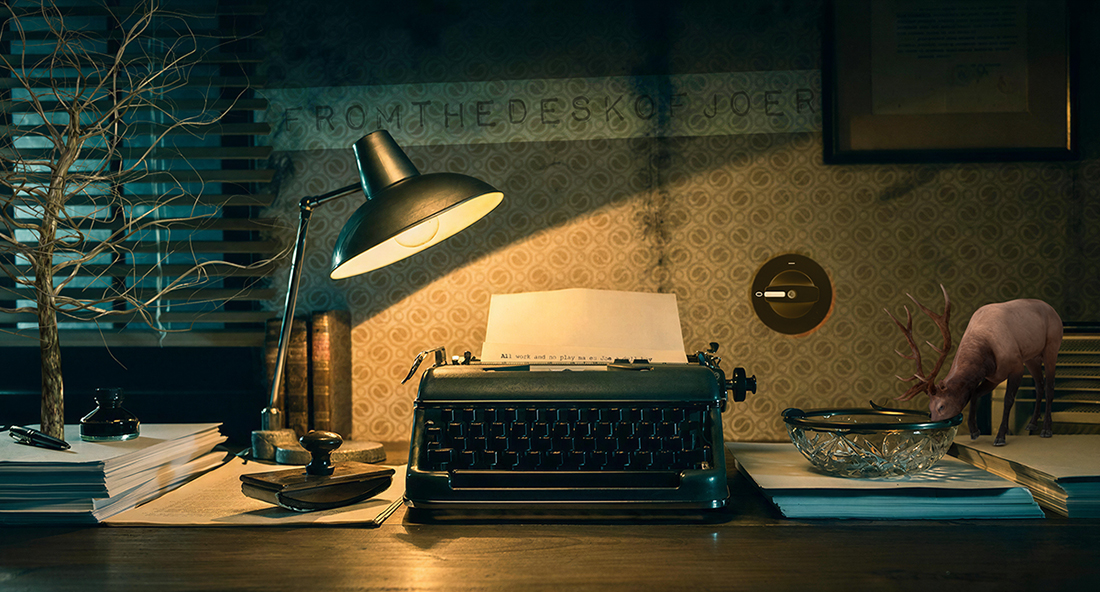 Semi-surreal noir office, a typewriter atop a desk stacked with tidy piles of paper. A leafless tree grows out of one pile; a tiny buck laps water out of a candy dish on another.
